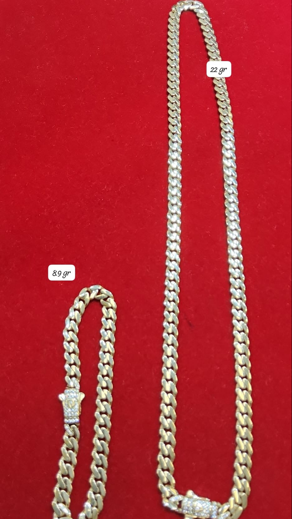 A gold bracelet and a gold necklace placed on a red surface, with the necklace appearing longer and the bracelet smaller in size.