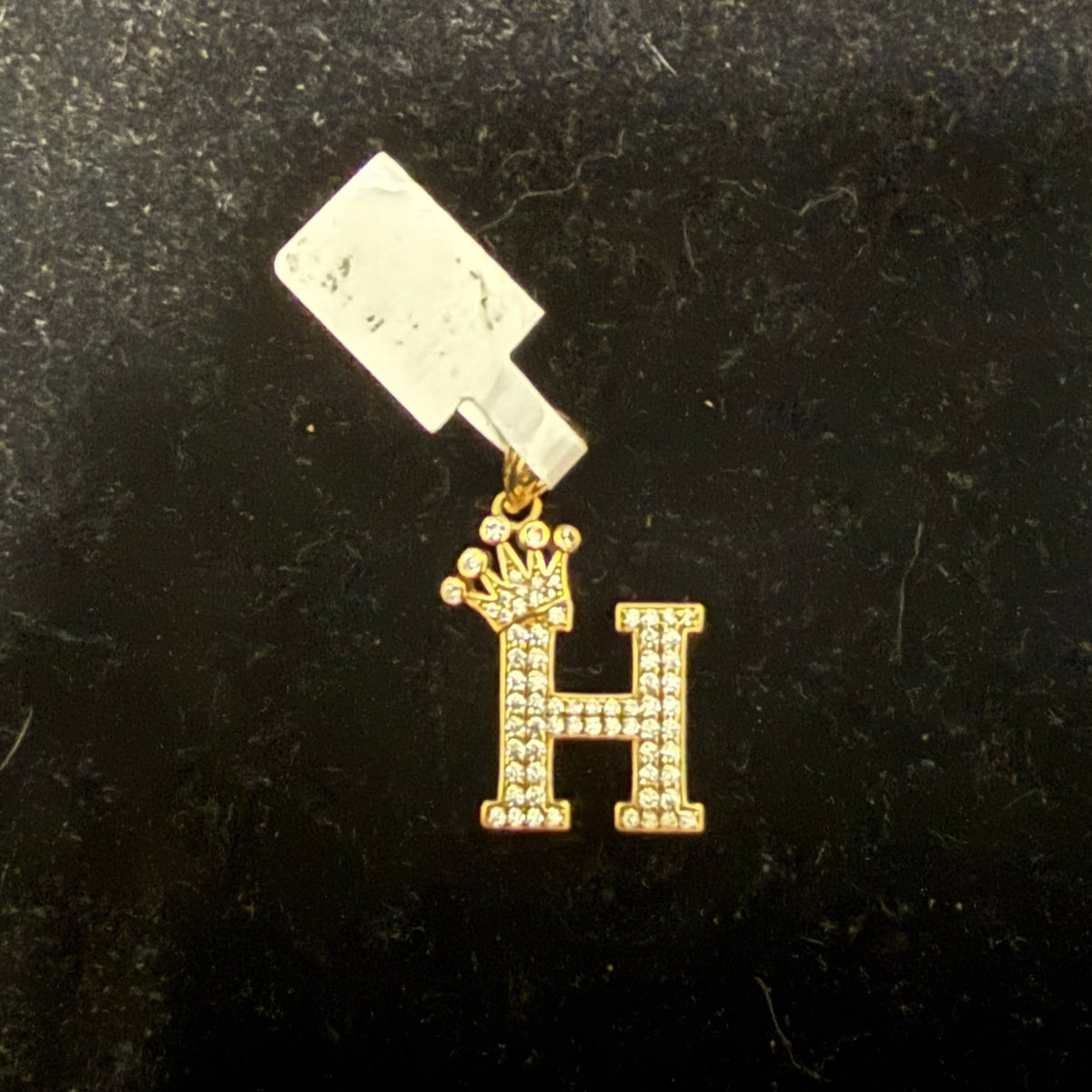 A gold-colored letter 'H' pendant with a clear crystal charm attached to it, displayed against a black background.