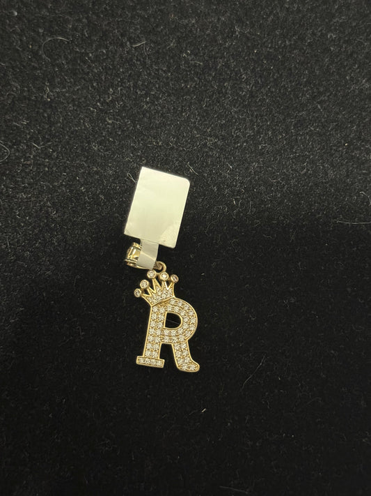 A gold-colored letter R charm with a crown on top, placed on a black background.